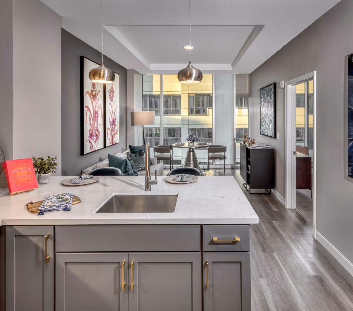 Modern senior living interior featuring a kitchen island with a sink and gray cabinetry, a dining area with a round glass table and chairs near large windows, and a living area with a sofa and decorative artwork on the walls. The space has pendant lighting and wood flooring.