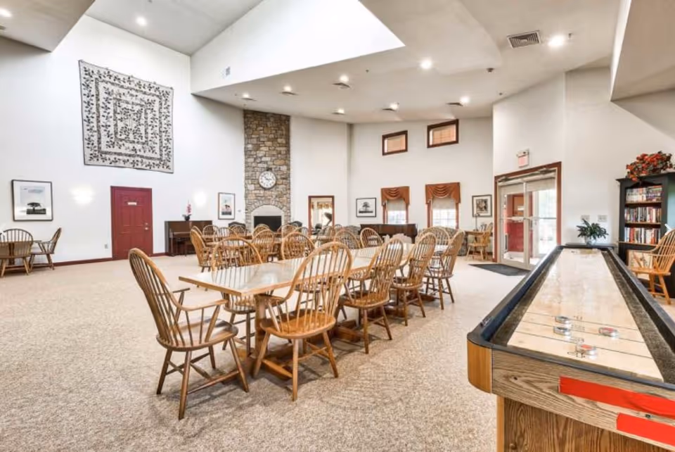 A spacious common area with multiple wooden tables and chairs arranged for group seating. The room features a stone fireplace with a clock above it, a piano, framed pictures on the walls, and a shuffleboard table in the foreground. There are windows with valances and a glass door leading outside. The ceiling has recessed lighting and a skylight.