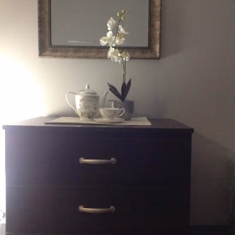 A dark wooden dresser with two drawers, topped with a white teapot, a matching teacup and saucer, and a potted white orchid. A framed mirror hangs on the wall above the dresser.
