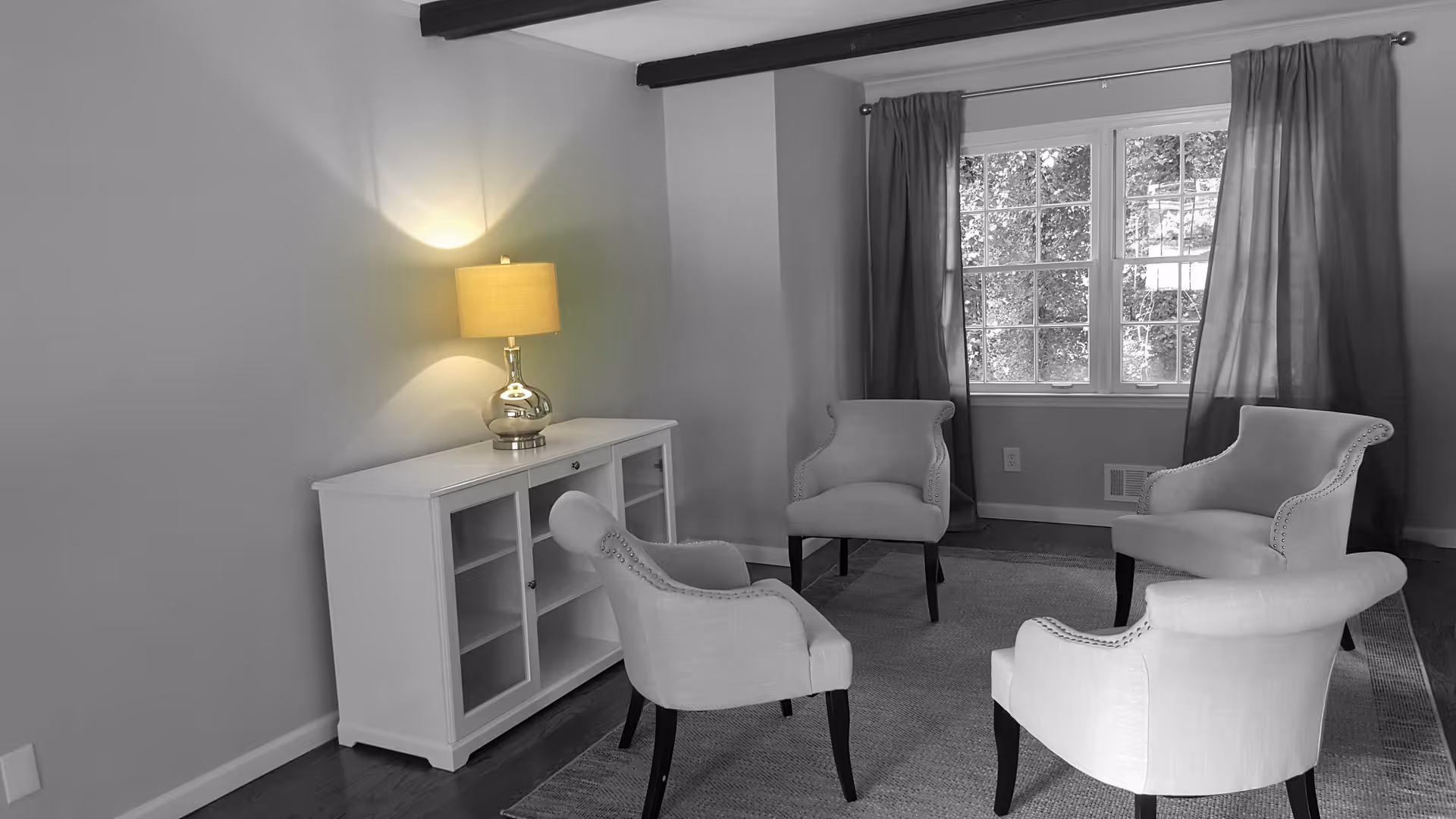 A cozy living room with four upholstered armchairs arranged in a circle on a rug. There is a white cabinet with glass doors against the wall, topped with a lit table lamp. A window with curtains lets in natural light.