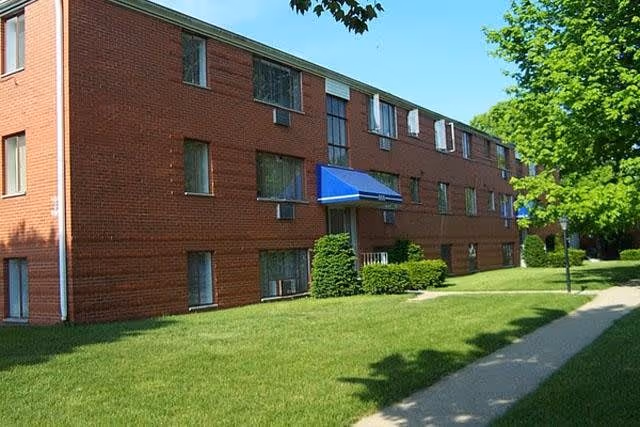 Exterior view of a three-story red brick apartment building with multiple windows and blue awnings over some entrances, surrounded by green grass, bushes, and trees under a clear blue sky.