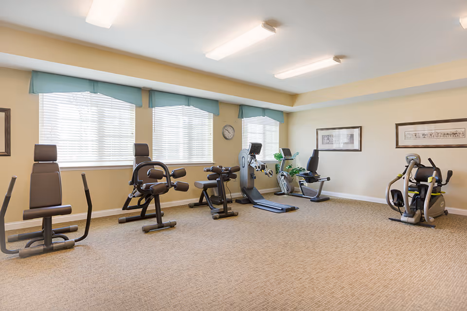 A well-lit exercise room with various fitness equipment including weight machines and stationary bikes, beige walls, carpeted floor, three windows with blue valances, and framed pictures on the walls.