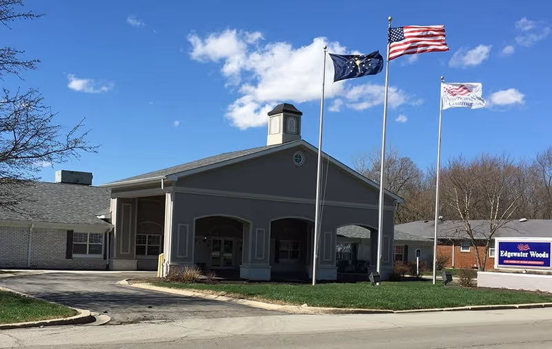 Exterior view of Edgewater Woods facility with a covered entrance, three flagpoles flying the American flag, a state flag, and a community flag, under a blue sky with some clouds.