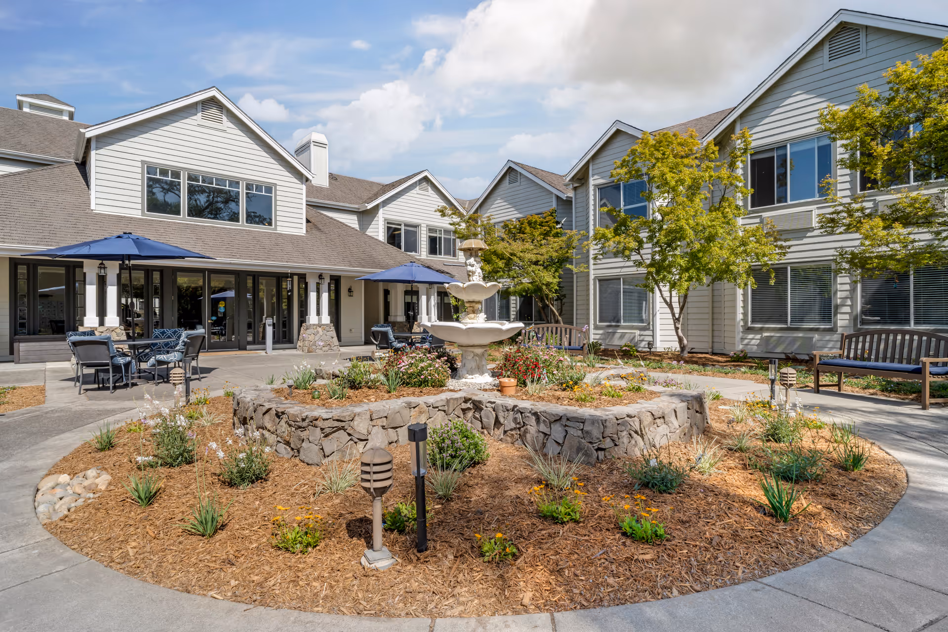 Courtyard with a central stone planter and fountain, outdoor seating and umbrellas in front of a two-story senior living building.