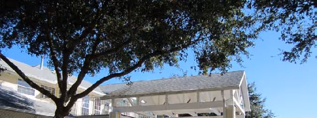 Outdoor view of a senior living facility with a large tree providing shade over a white pergola structure. The building has white siding and multiple windows under a clear blue sky.