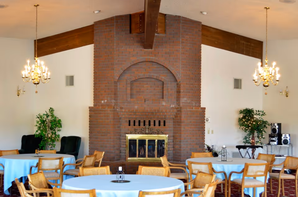 A spacious common area with round tables covered in light blue tablecloths surrounded by wooden chairs. The room features a large brick fireplace with a brass screen in the center, two chandeliers hanging from the ceiling, and green potted plants on either side. There is also a stereo system on a small table to the right.