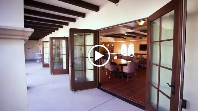 View of an open corridor with multiple glass-paneled wooden doors leading into a dining area furnished with round tables and chairs. The corridor has a light-colored floor and ceiling with exposed wooden beams, and the dining area features arched windows and warm lighting.