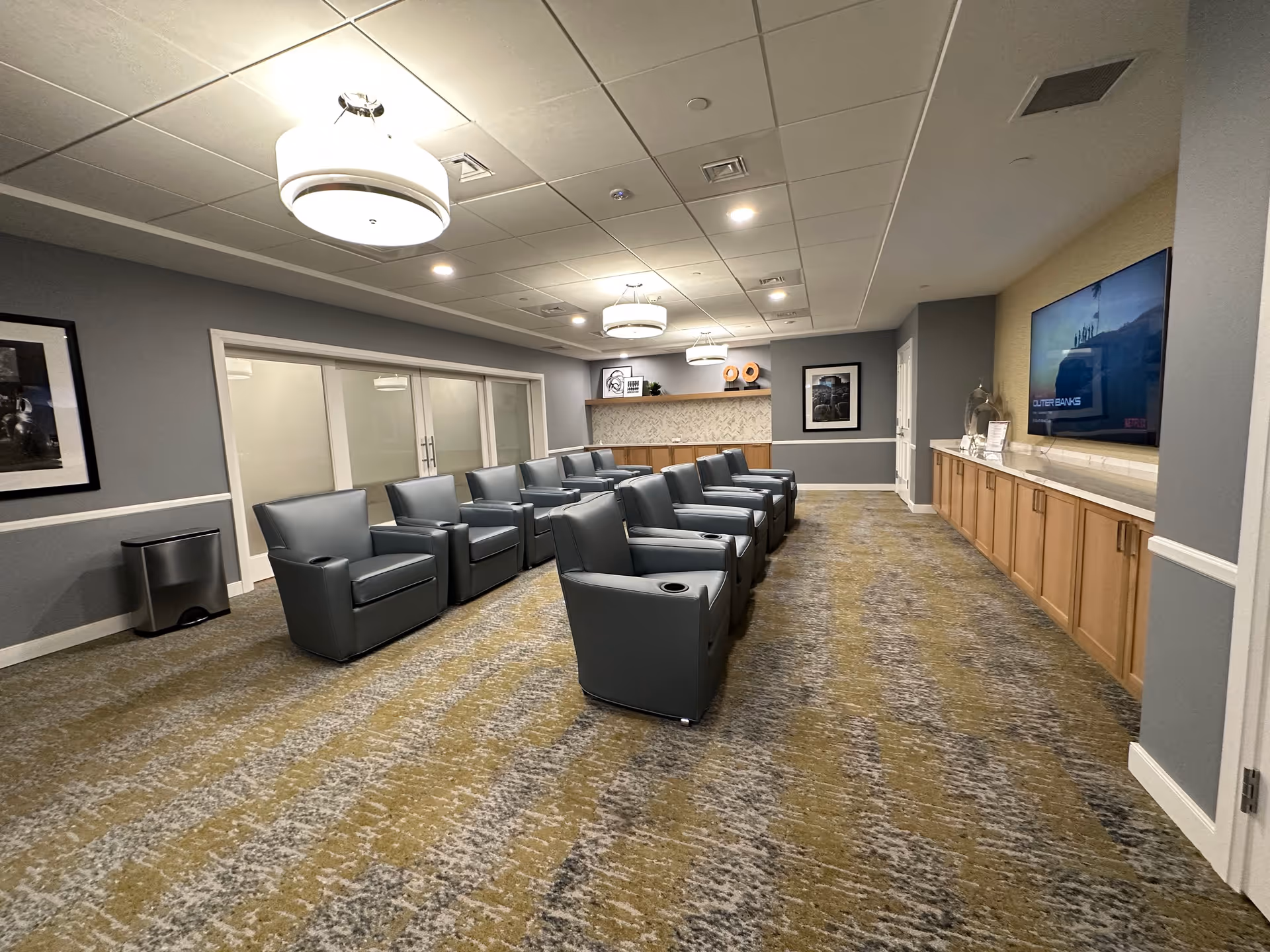 A spacious room with two rows of gray leather recliner chairs facing a large flat-screen TV mounted on the wall. The room has a patterned carpet in shades of yellow and gray, gray walls with white trim, and modern ceiling lights. There are framed pictures on the walls and wooden cabinets beneath the TV and along the back wall.