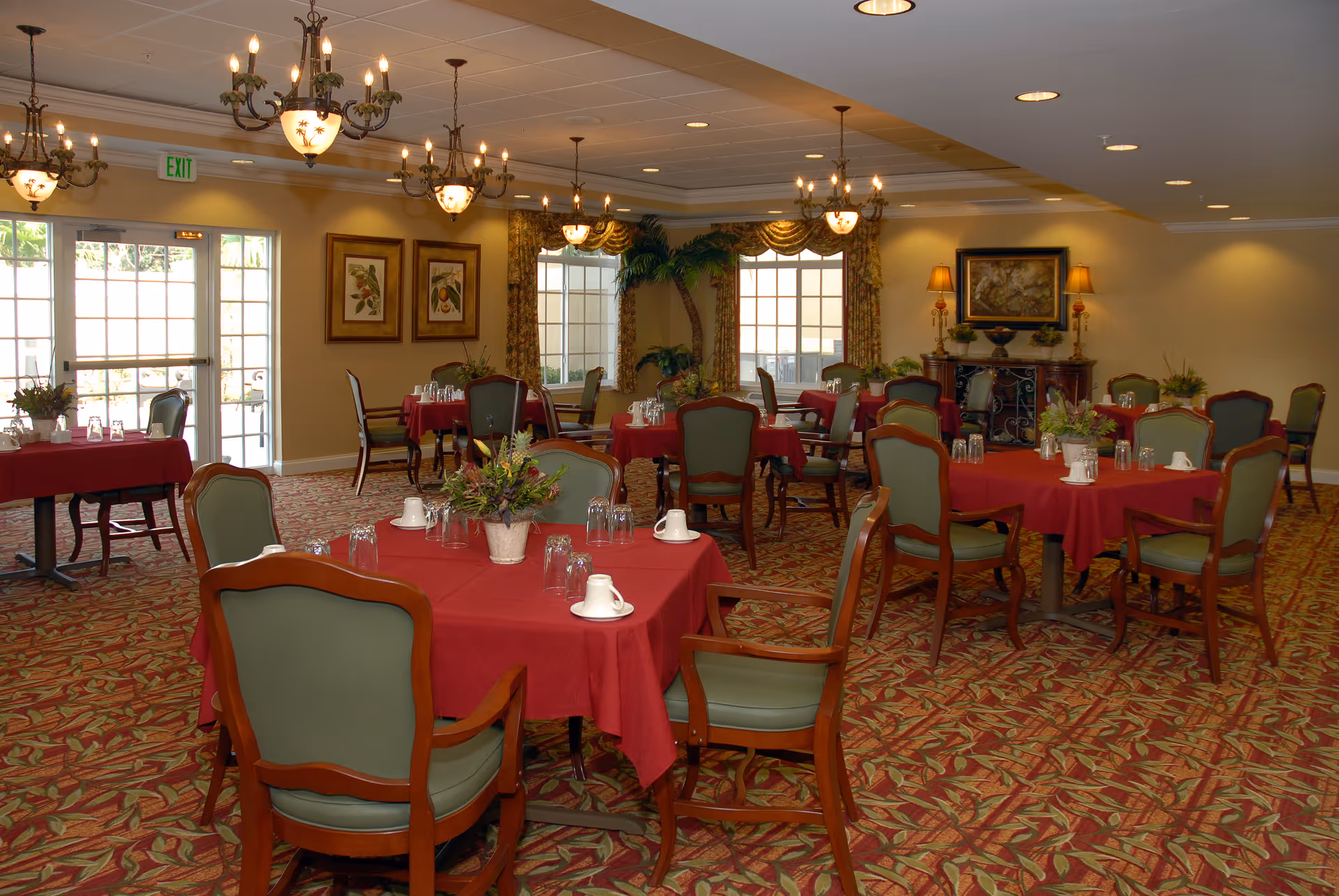 A dining room with multiple round tables covered in red tablecloths, each set with cups, glasses, and floral centerpieces. The room has green cushioned wooden chairs, patterned carpet, chandeliers hanging from the ceiling, large windows with curtains, framed artwork on the walls, and a sideboard with lamps and a painting.