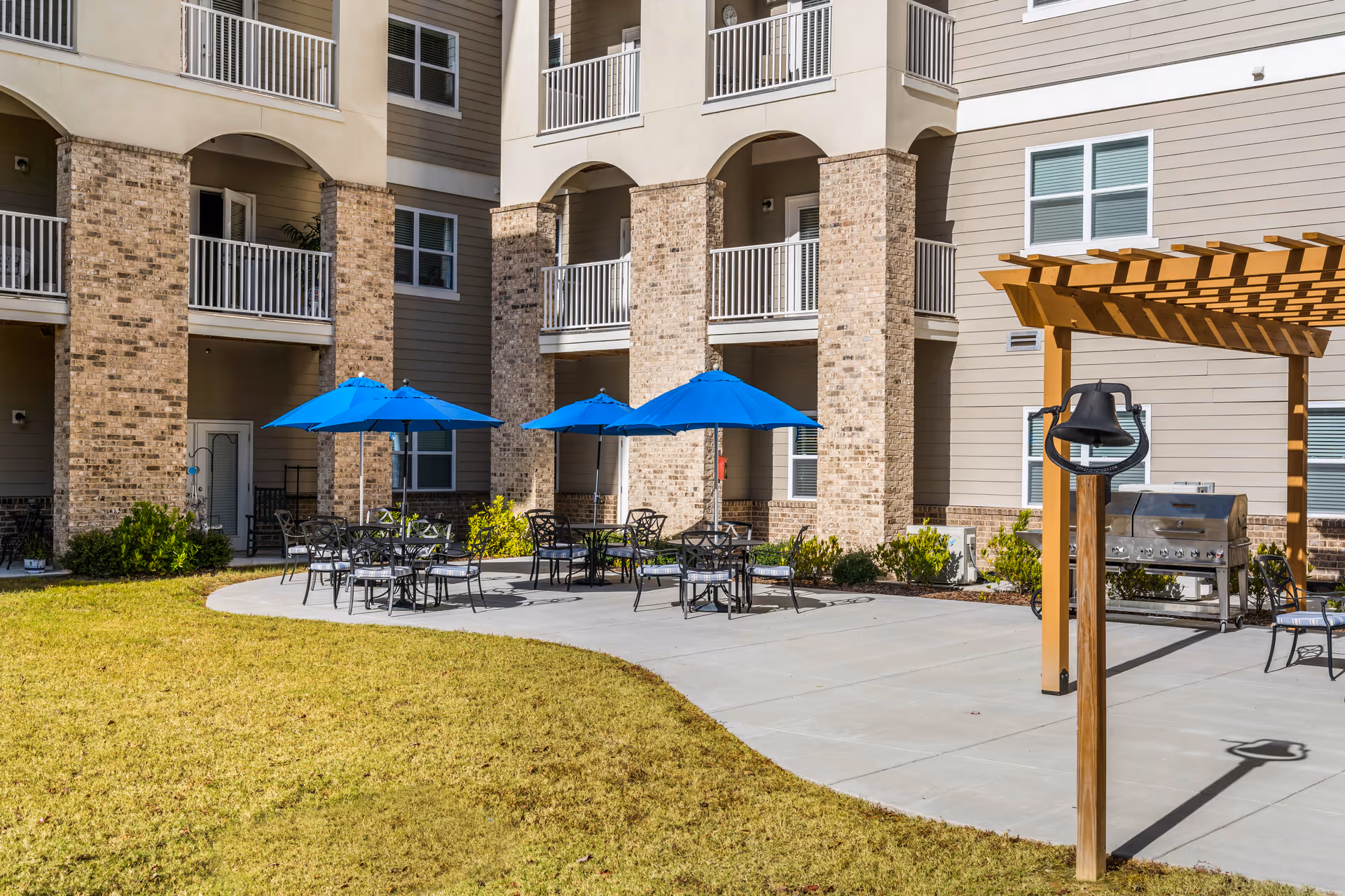 Outdoor patio area at The Mansions at Gwinnett Park Senior Independent Living featuring several black metal tables and chairs with blue umbrellas, a wooden pergola with a mounted bell, a barbecue grill, and a multi-story building with balconies in the background.