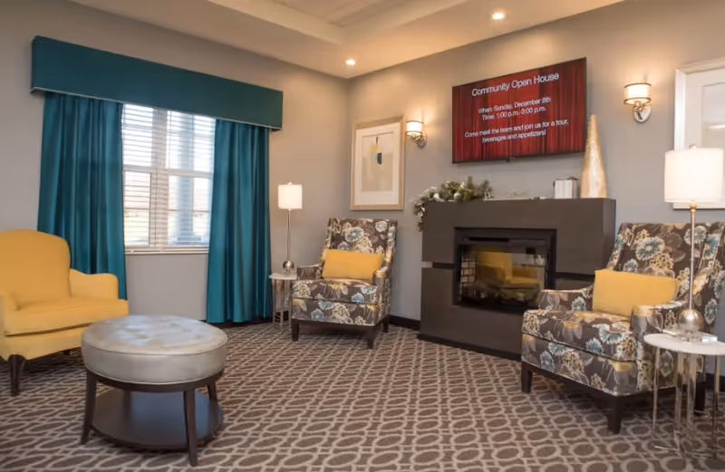 Cozy communal seating area with patterned armchairs, yellow accent chairs, a fireplace, wall-mounted TV, and teal curtains.