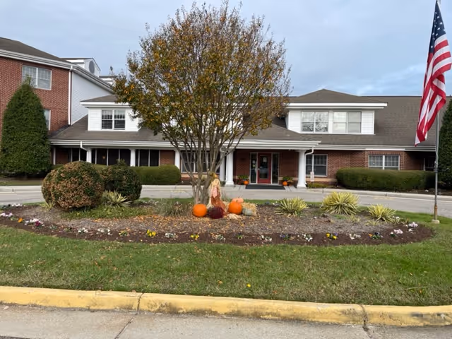 Brick senior living building with manicured landscaping, pumpkins, a tree, and an American flag at entrance.