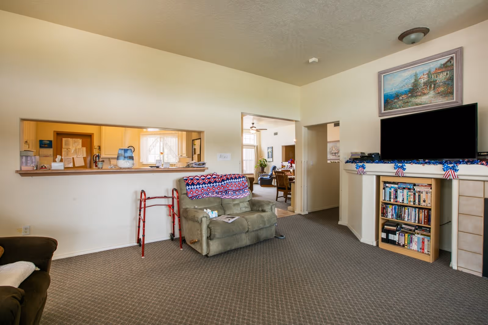 A cozy living room in an assisted living facility with a green recliner chair covered by a patterned blanket, a red walker beside it, a TV mounted above a fireplace decorated with blue and red garland, and a bookshelf filled with DVDs and books. The room has beige walls and carpeted flooring, with a pass-through window to the kitchen and a view into a dining area in the background.