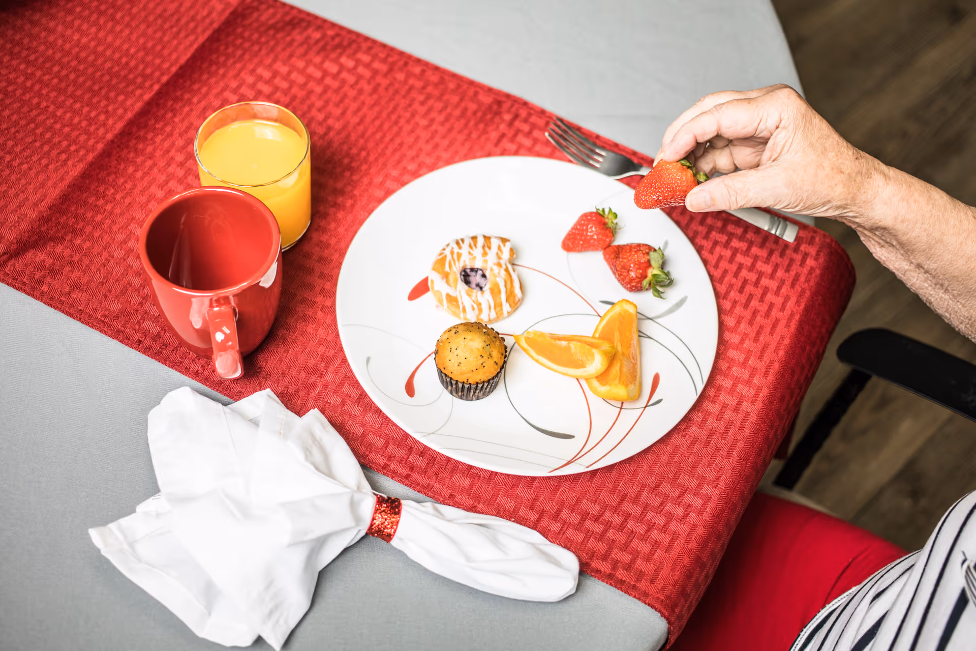 A close-up of a table set with a red table runner and a gray tablecloth. On the table is a white plate with a muffin, a pastry with icing, orange slices, and strawberries. A hand is reaching to pick up a strawberry. There is also a red mug, a glass of orange juice, a fork, and a folded white napkin with a red ring on the table.