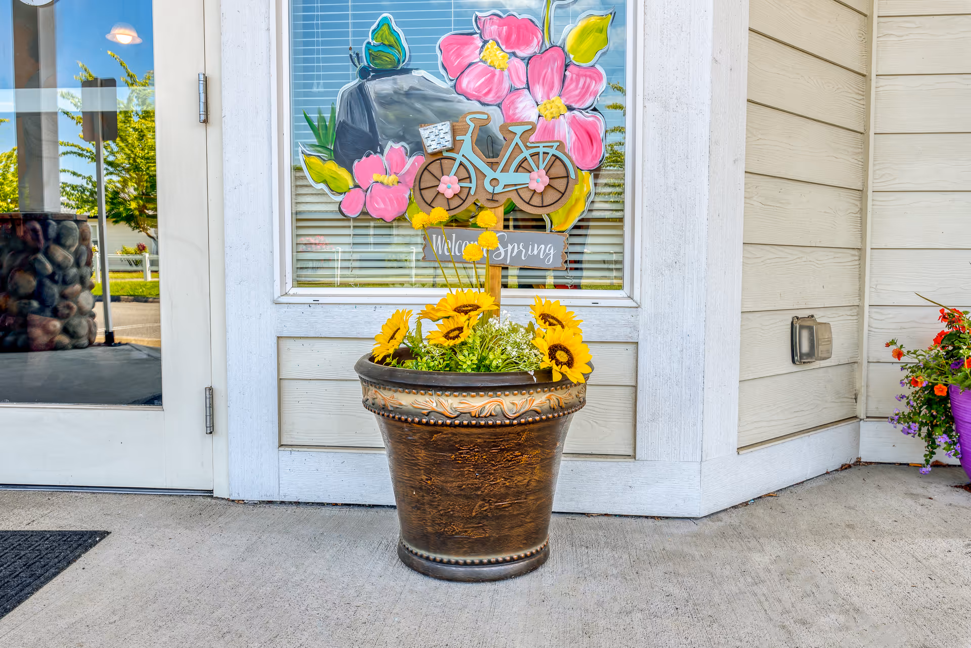 A large decorative flower pot with sunflowers and other greenery placed outside a building entrance. Behind the pot is a window with a colorful spring-themed decoration featuring a bicycle, pink flowers, and a butterfly, along with a sign that says 'Welcome Spring'. The building exterior has light-colored siding and a glass door to the left.