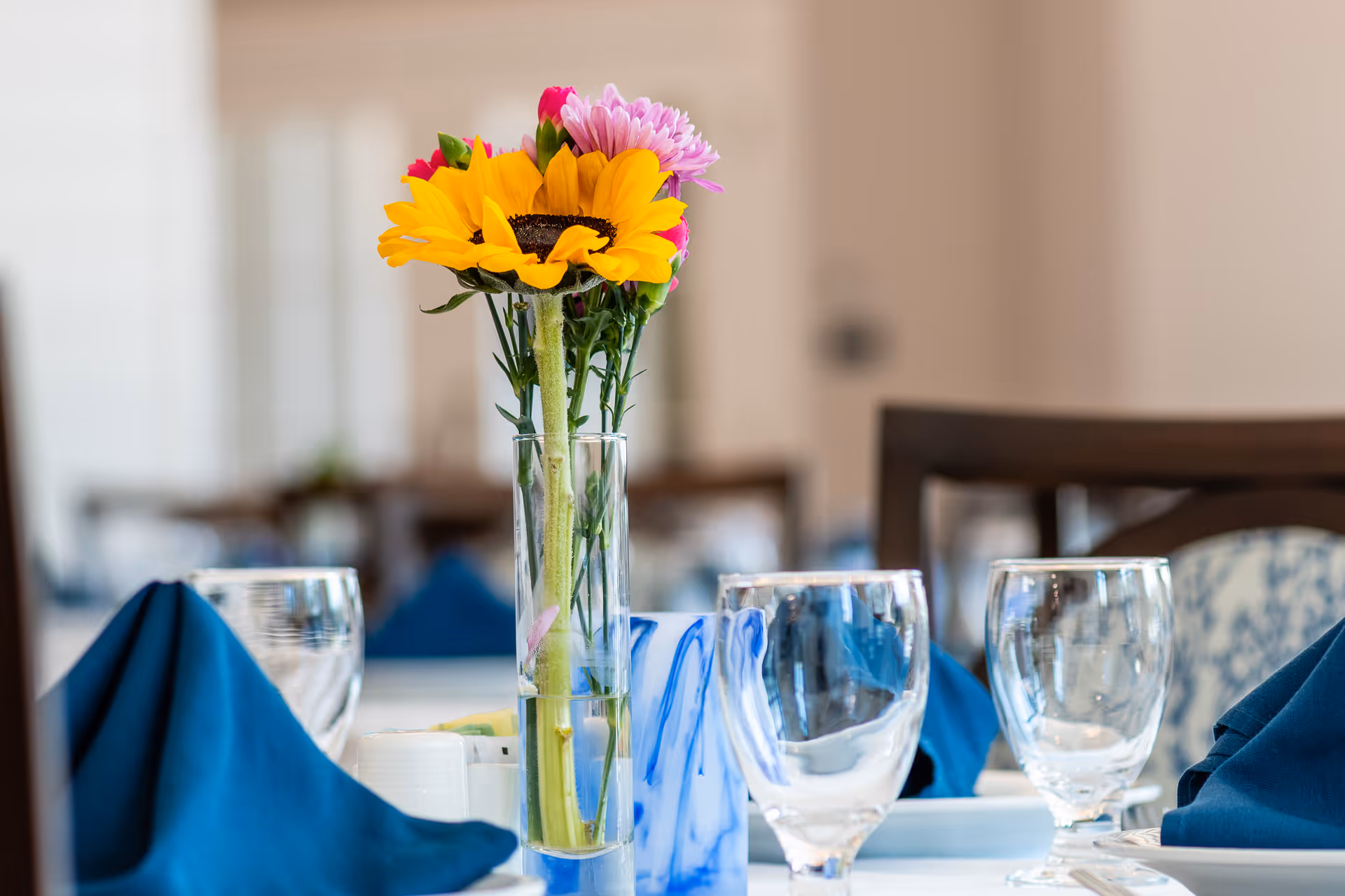 Close-up of a dining table set with clear water glasses, blue folded napkins, and a tall glass vase holding a bouquet of colorful flowers including a sunflower and pink blooms, in a softly lit dining room.