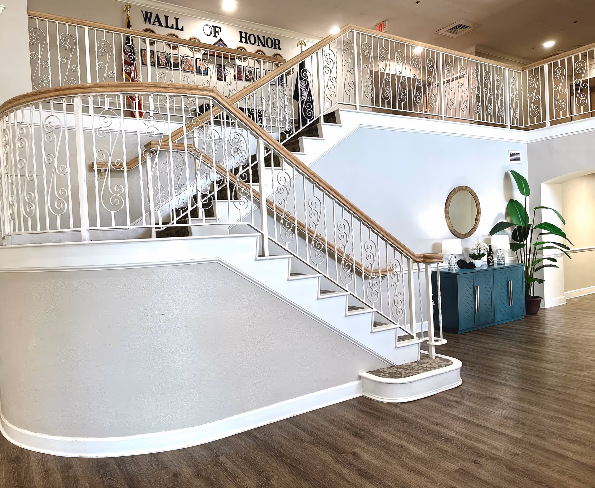 Interior view of a senior living facility showing a decorative staircase with white railings and wooden handrails. On the wall above the stairs is a 'Wall of Honor' display with photos and plaques. Below the stairs is a teal cabinet with lamps, plants, and a round mirror above it. The floor is wood-style, and the walls are painted light gray.
