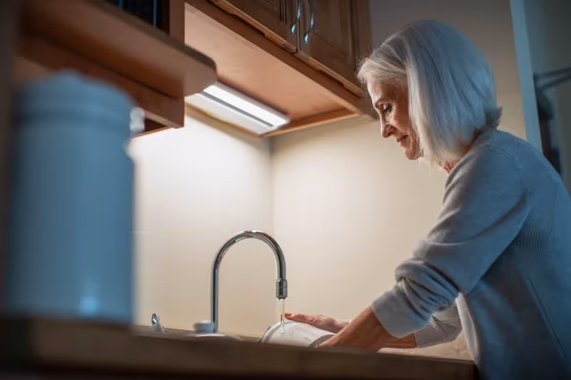 An older woman washing dishes at a kitchen sink under under-cabinet lighting.