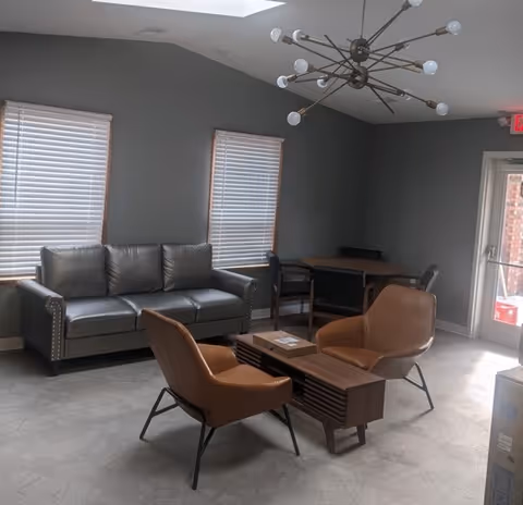 A modern living room with a gray leather sofa against a gray wall with two windows covered by white blinds. In front of the sofa are two brown leather chairs and a wooden coffee table. A round wooden table with four chairs is in the corner near a glass door. A contemporary chandelier with multiple light bulbs hangs from the ceiling.