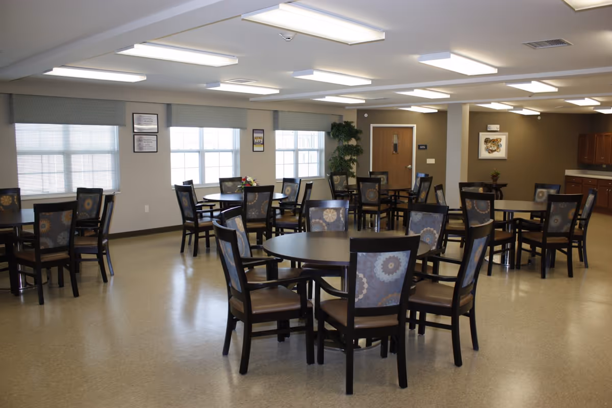 A spacious dining room with multiple round tables and chairs arranged neatly. The chairs have patterned backs and cushioned seats. Large windows with blinds allow natural light to fill the room. The walls are decorated with framed pictures and a potted plant is placed near a door. The ceiling has several rectangular fluorescent lights.