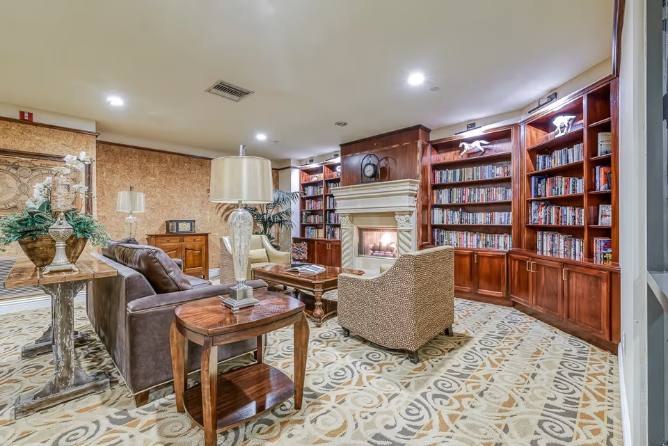 A cozy living room area with a fireplace surrounded by built-in wooden bookshelves filled with books. The room features a patterned carpet, a brown leather sofa, a beige armchair, wooden side tables with lamps, and decorative plants and ornaments. The lighting is warm and inviting.