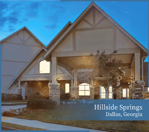 Exterior view of a senior living facility building with stone pillars and a peaked roof at dusk, with lights illuminating the entrance area.