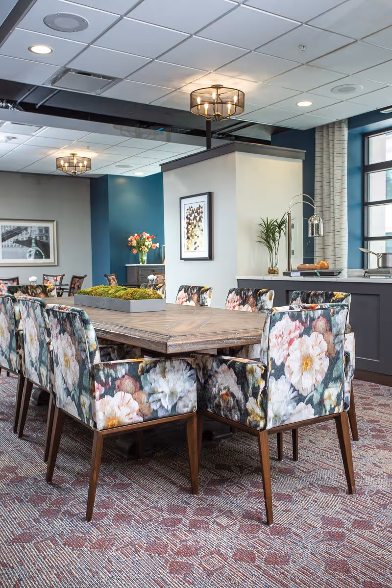 Communal dining area with a wooden table surrounded by floral-upholstered chairs, decorative lighting, and a service counter in the background.