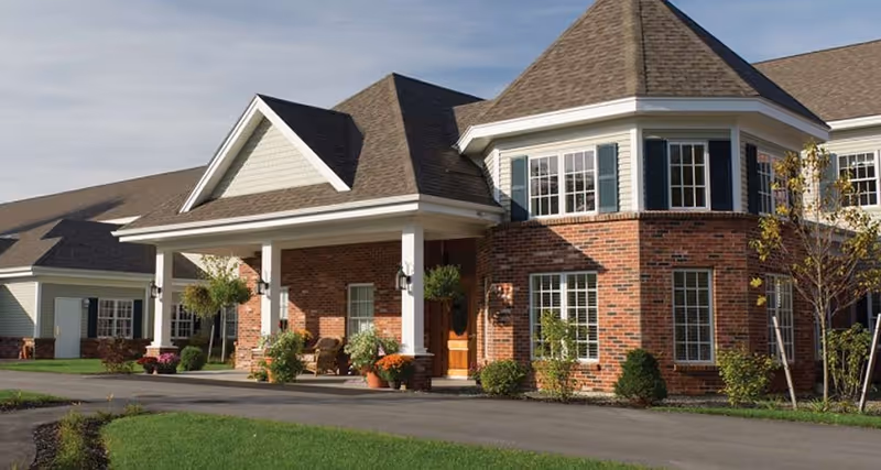 Exterior view of a senior living community building with brick and siding walls, multiple windows, a covered entrance with white columns, and landscaping including bushes and small trees under a partly cloudy sky.