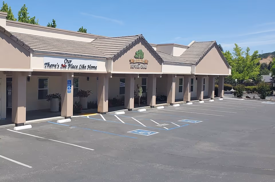 Front exterior of Village Oaks Senior Care showing the entrance with a covered walkway, signage, and empty parking spaces including accessible spots.