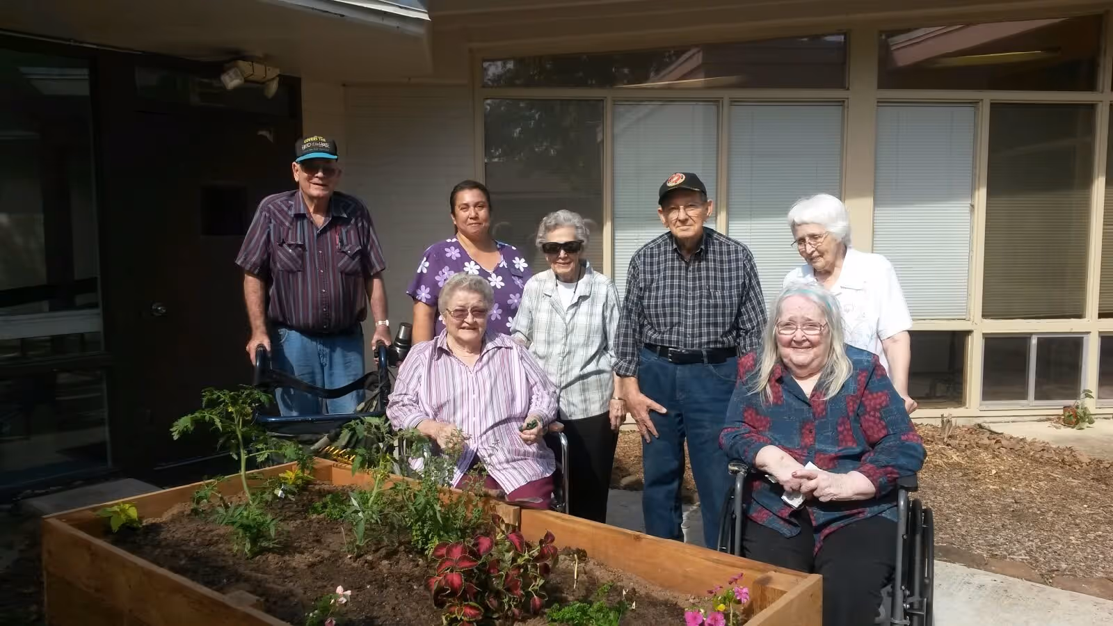 A group of elderly people and a caregiver standing and sitting around a raised garden bed outside a building. The garden bed contains various plants and flowers. Two elderly women are in wheelchairs, and the group is smiling and enjoying the outdoor setting.
