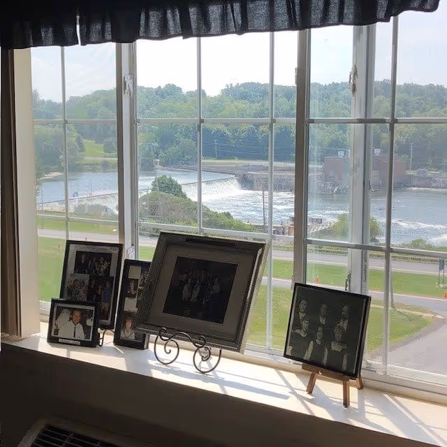 A window with a black valance at the top overlooks a river with a small dam and greenery in the background. On the windowsill, there are several framed photographs of people arranged in a row.