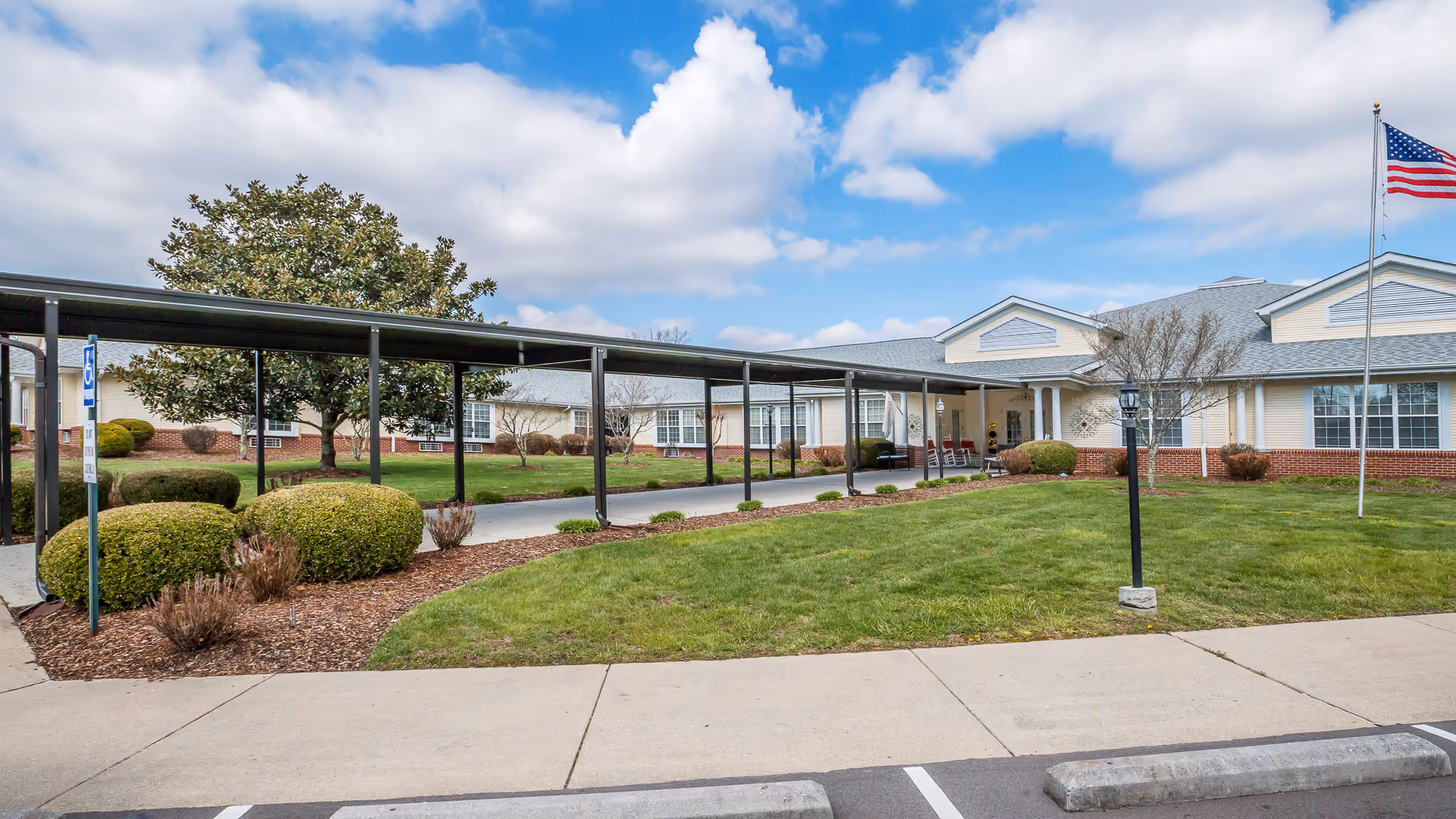 Front exterior view of a single-story senior living facility building with a covered walkway, manicured bushes, green lawn, and an American flag on a flagpole under a partly cloudy sky.