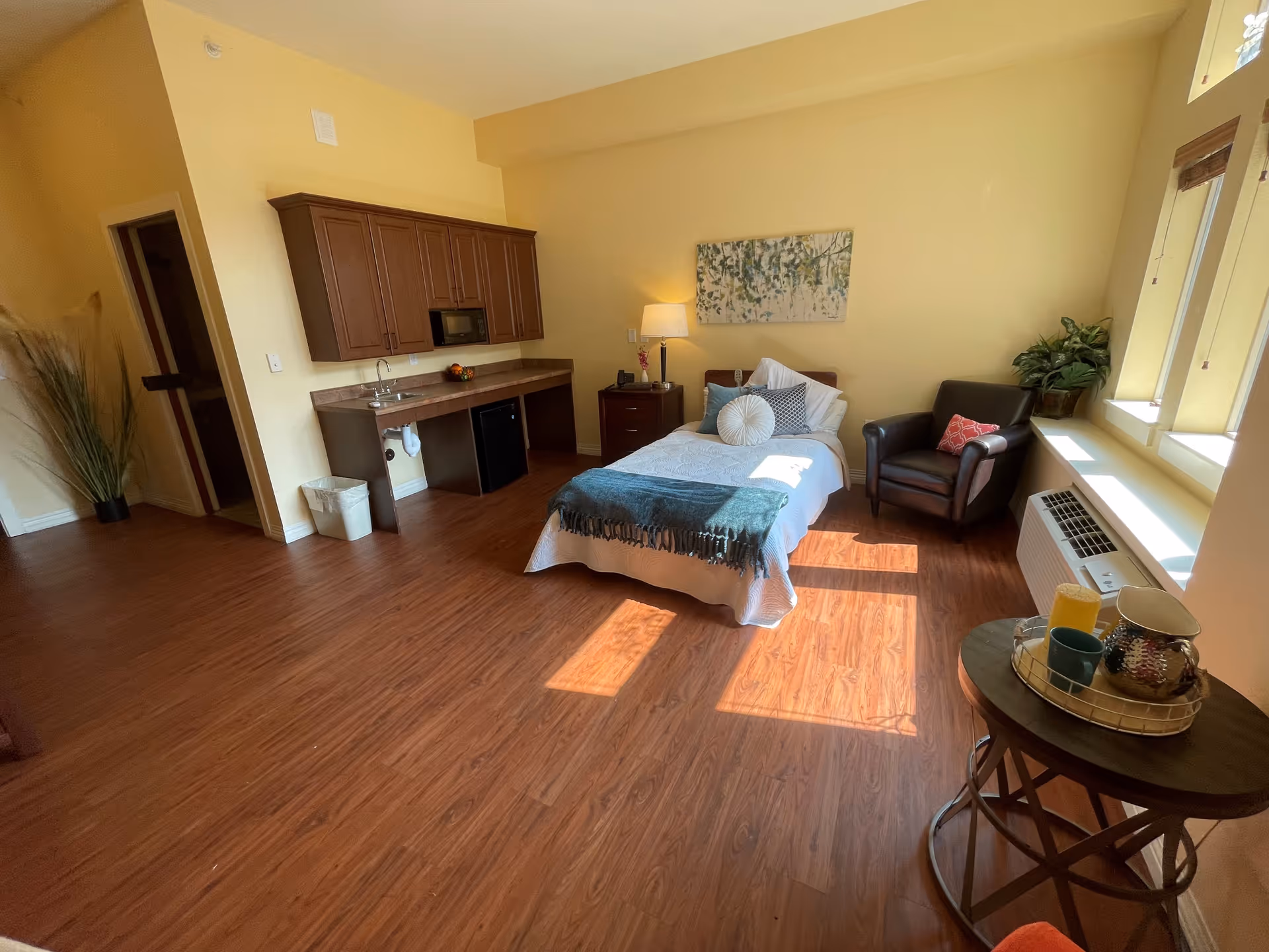 A bright, cozy room with a single bed adorned with pillows and a throw blanket. There is a dark brown armchair with a red patterned pillow next to a window with blinds. A small round table holds a tray with a pitcher, cups, and a candle. On the left side, there is a kitchenette with wooden cabinets, a sink, a microwave, and a mini fridge. The floor is wood, and the walls are painted light yellow. A tall plant is placed near the doorway.