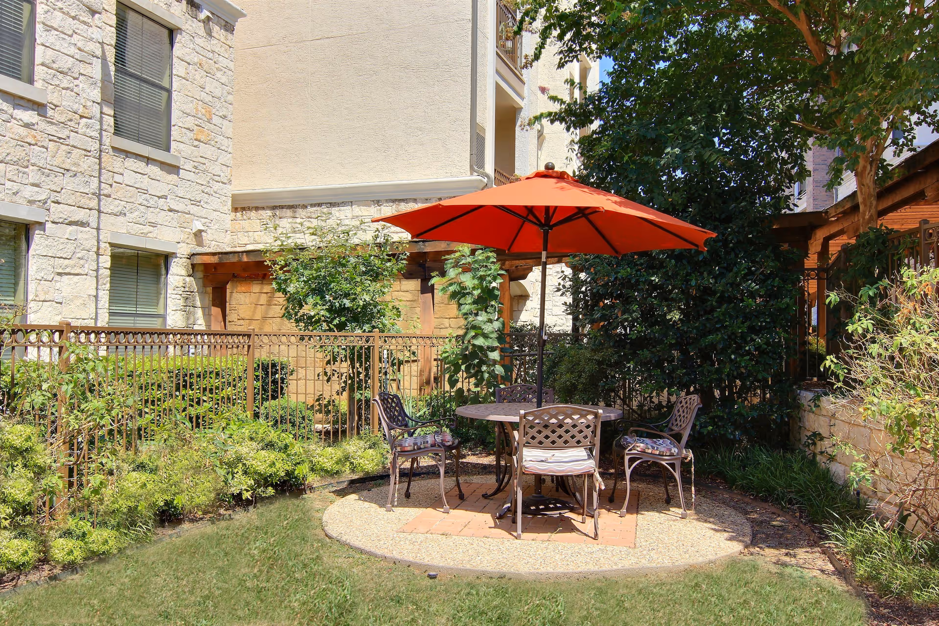 Outdoor seating area with a round metal table and four metal chairs with cushions, shaded by a large orange umbrella. The area is surrounded by greenery, including bushes and trees, and is adjacent to a building with stone and stucco walls.