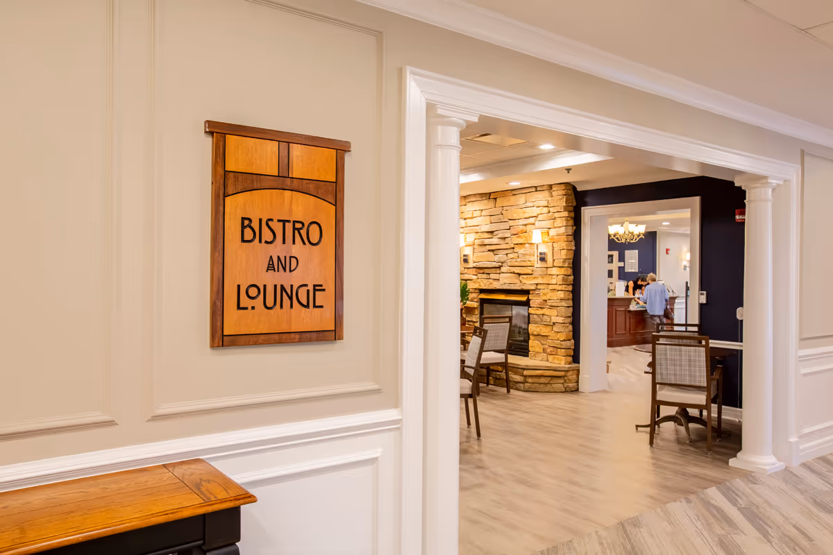 Interior view of a senior living facility hallway leading to a bistro and lounge area. The hallway features white walls with decorative molding and a wooden sign that reads 'Bistro and Lounge'. Beyond the hallway, there is a cozy lounge area with a stone fireplace, chairs, and warm lighting. In the background, a few people are seen near a reception or service desk.