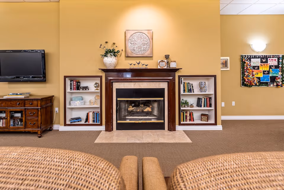 Comfortable senior living common room with a central fireplace flanked by built-in bookshelves, a wall-mounted TV, and seating.