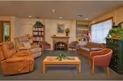 A cozy senior living common area with comfortable seating including a brown recliner, beige armchair, patterned sofa, and a red armchair arranged around a wooden coffee table. There is a fireplace with a painting above it, bookshelves on either side, and a large window with blinds letting in natural light. A green chest of drawers with a plant on top is visible on the right side.