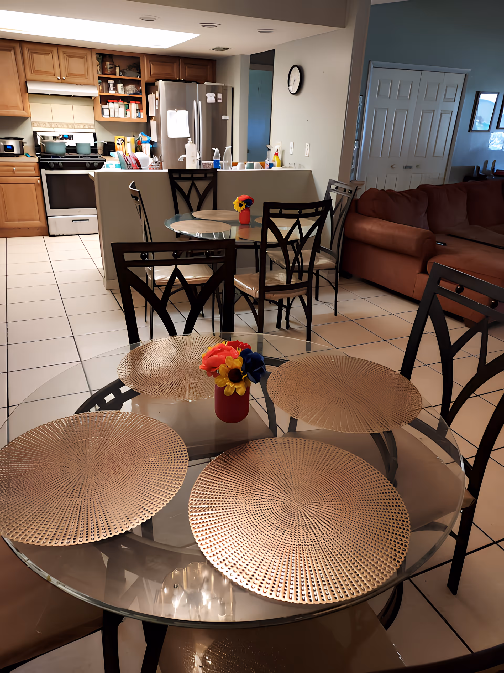 Open-concept interior with glass dining tables and chairs in the foreground, a kitchen with stove and refrigerator in the background, and a sofa to the right.