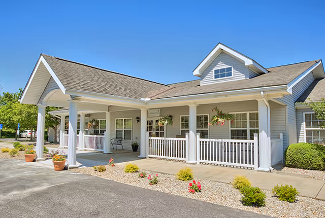 Exterior view of a single-story building with a covered porch, white railings, and hanging flower baskets under a clear blue sky. The building has a gabled roof and is surrounded by small shrubs and flower beds.