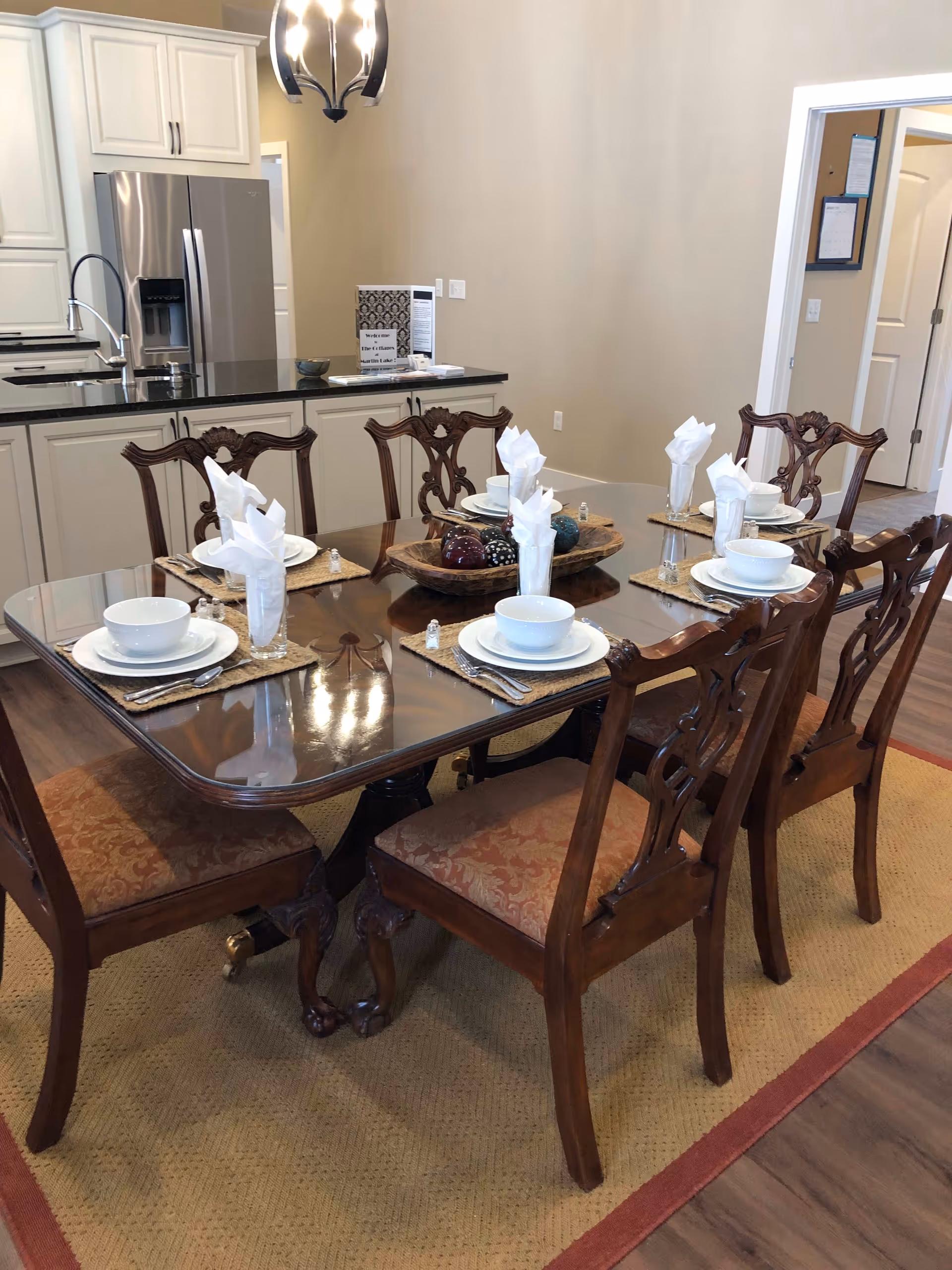A dining area with a polished wooden table set for six people. Each place setting includes a white bowl, plate, glass with a folded white napkin, and silverware on woven placemats. The table is surrounded by six ornate wooden chairs with cushioned seats. In the background, there is a kitchen area with white cabinets, a stainless steel refrigerator, and a black countertop. A decorative light fixture hangs above the table.