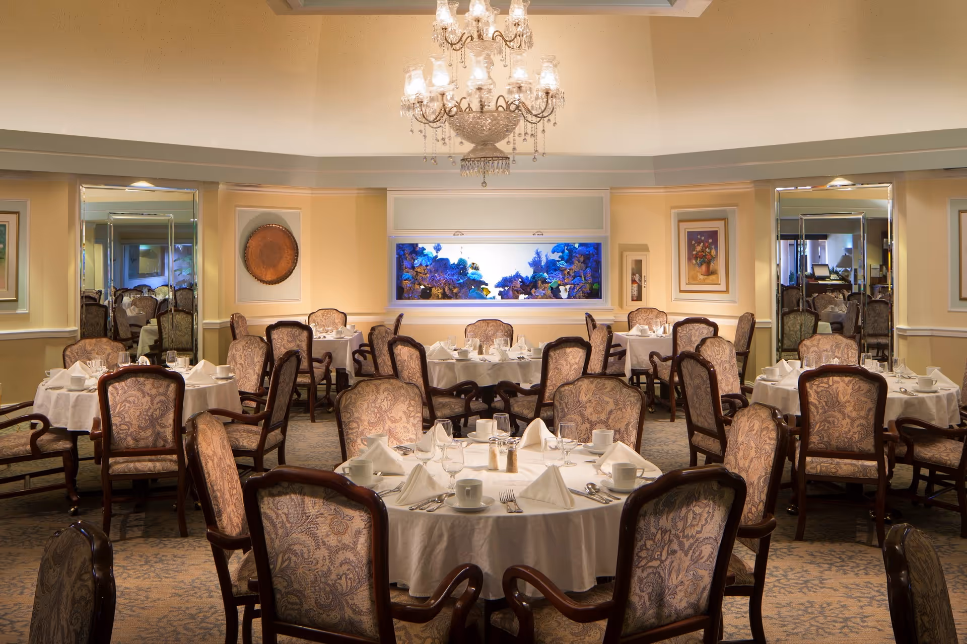 Elegant dining room with round tables covered in white tablecloths, set with white napkins, cups, and silverware. The room features upholstered chairs with ornate patterns, a large chandelier hanging from the ceiling, framed artwork on the walls, and a large built-in aquarium filled with colorful coral and fish.