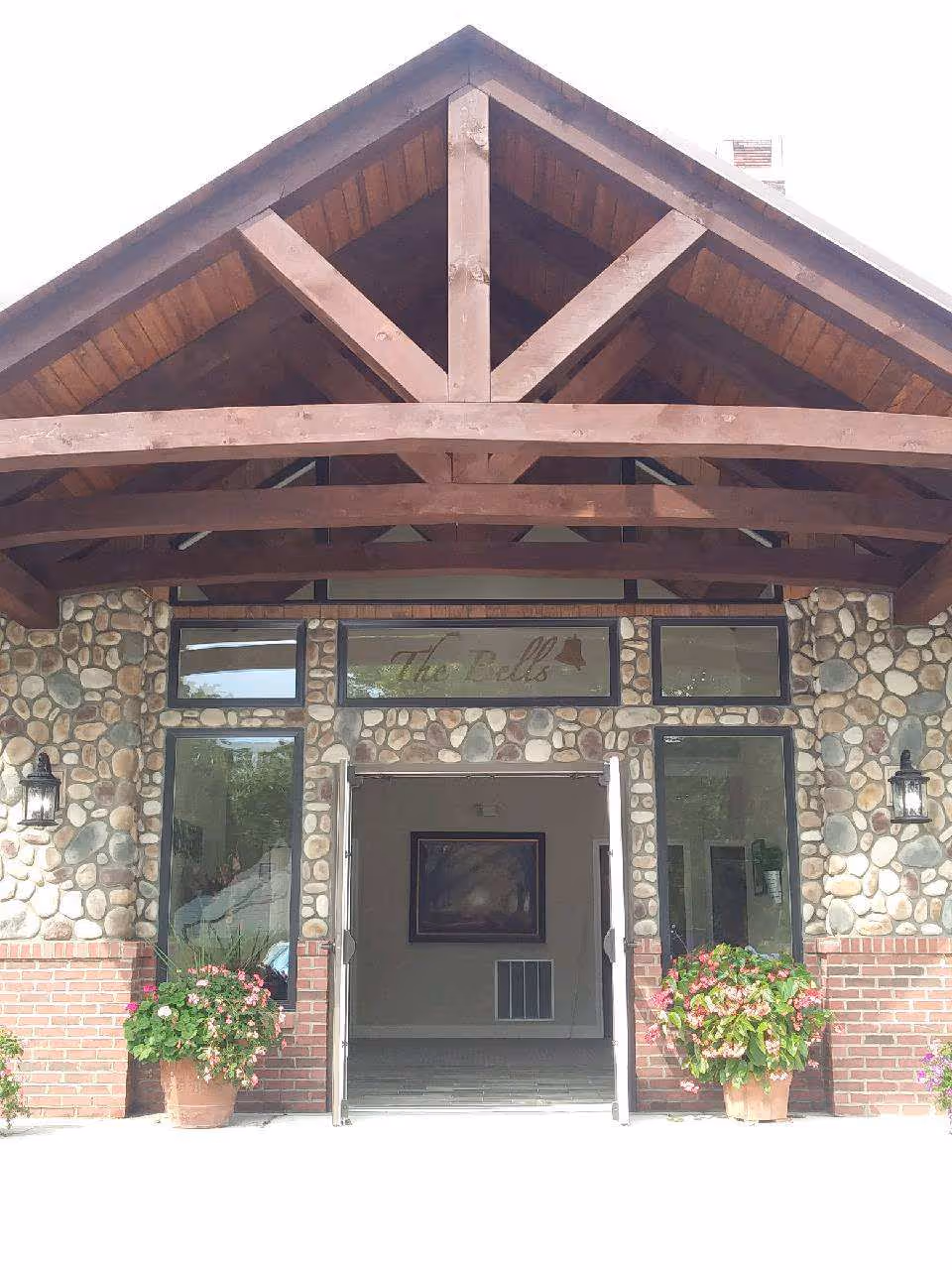 Front entrance of a stone-and-brick building with a large wooden gabled canopy, open double doors, and potted flower planters.