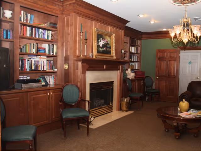 A cozy living room with wooden built-in bookshelves filled with books, a fireplace with a painting above it, green upholstered chairs, a wooden coffee table with magazines and decorative items, and a chandelier hanging from the ceiling.