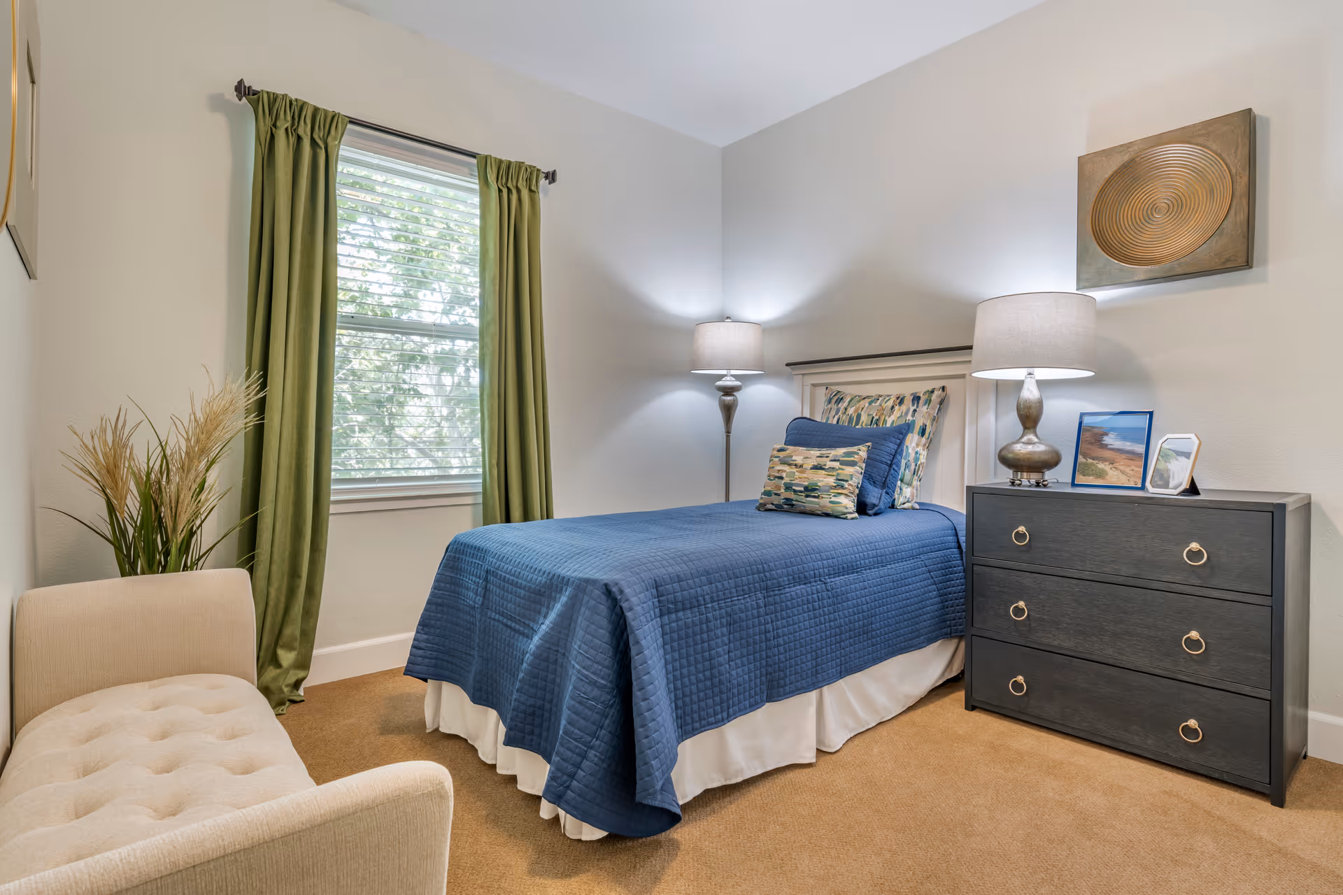 A cozy bedroom with a single bed covered in a blue quilt and decorative pillows. Next to the bed is a dark wooden dresser with three drawers, topped with a lamp and framed pictures. A beige upholstered bench is placed near a window with green curtains, and a decorative plant is beside the bench. The walls are light-colored, and a piece of circular wall art hangs above the dresser.