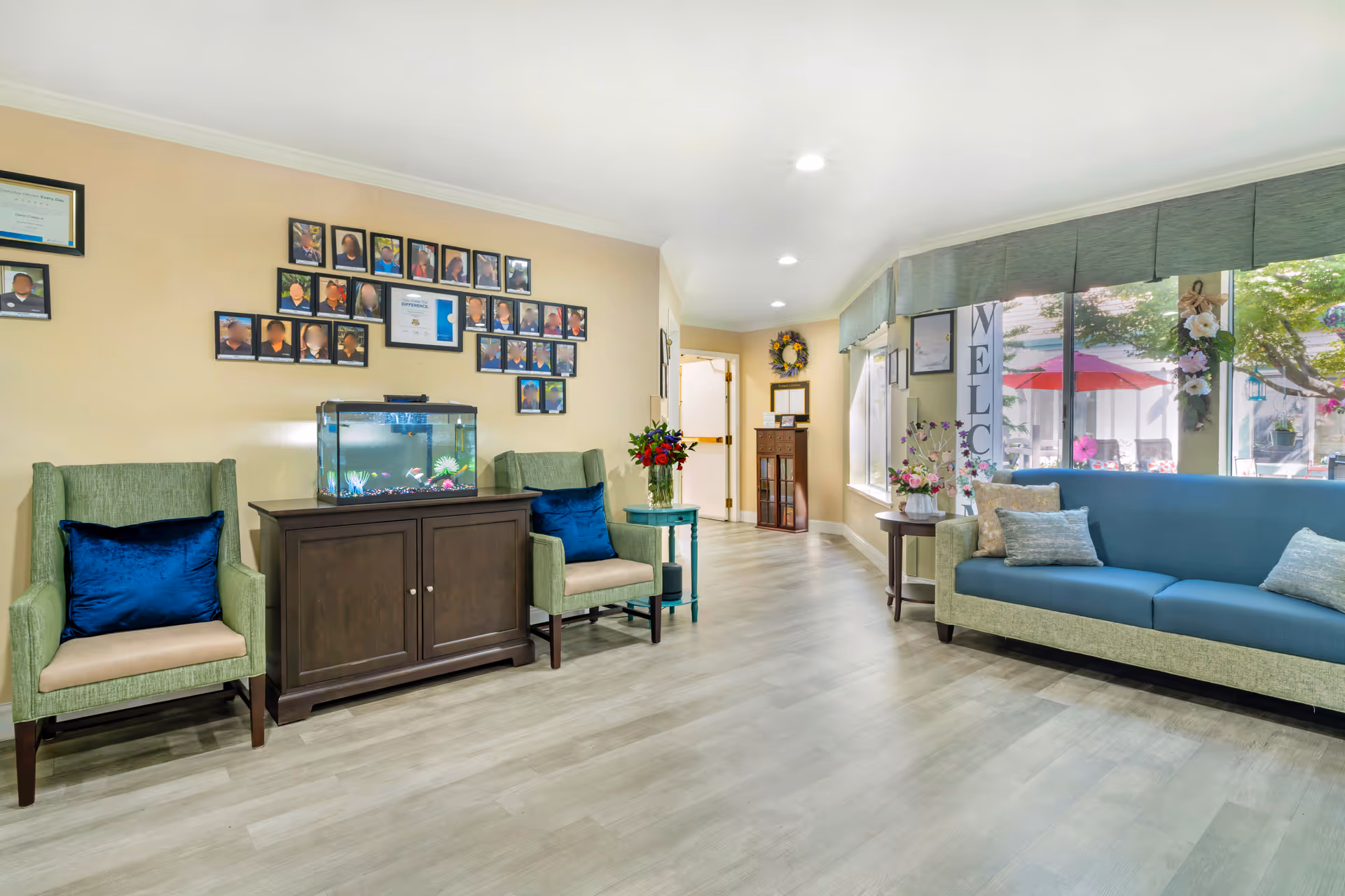 Bright reception lounge with armchairs and a sofa, an aquarium on a cabinet, framed photos on the wall, and large windows looking out to an outdoor patio.