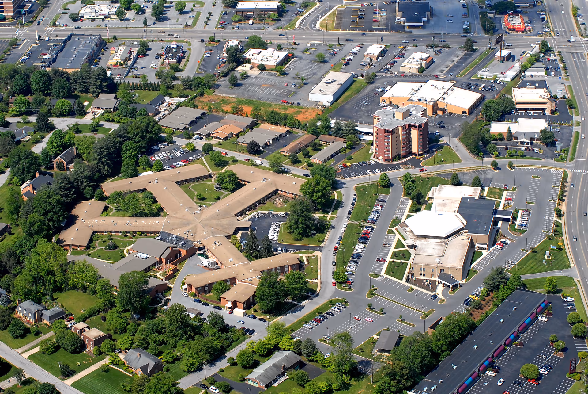 Aerial view of Abundant Christian Living Community showing multiple buildings arranged with parking lots, roads, and green spaces surrounding the facility. The main building has a star-like layout with several wings extending outward, surrounded by trees and other smaller buildings nearby.