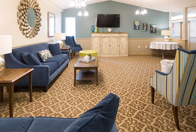 Spacious communal living room with blue sofas, patterned carpet, a wall-mounted TV above a cabinet, and accent chairs.