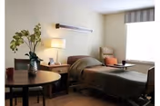 A well-lit patient room with a single hospital bed covered in a brown blanket, a wooden nightstand with a lamp and picture frame, a comfortable armchair with an orange cushion near a large window with striped valance, and a round wooden table with a vase of flowers and a cup.