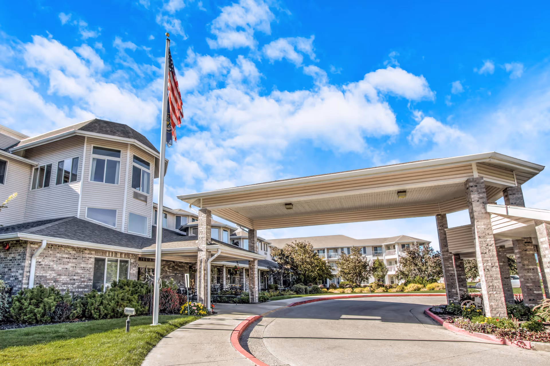 Exterior view of Pioneer Valley Lodge Gracious Retirement Living showing the entrance with a covered driveway supported by brick pillars, an American flag on a flagpole, and a multi-story building with windows under a partly cloudy blue sky.