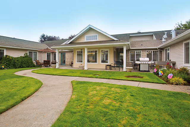 Exterior view of a single-story residential building with beige siding and a covered porch. The building is surrounded by a well-maintained green lawn and a curved concrete walkway. Outdoor seating and a barbecue grill are visible on the porch and patio areas.